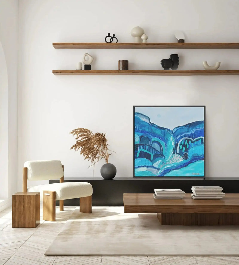 Large framed canvas art print displayed on a black console table in a minimalist living room. The abstract painting features a landscape of rolling hills and architectural elements in various shades of blue and teal, with a black frame. The room includes a cream upholstered armchair, two wooden side tables, a wooden coffee table with books, a grey spherical vase with dried pampas grass, and a cream rug on a light wood floor.