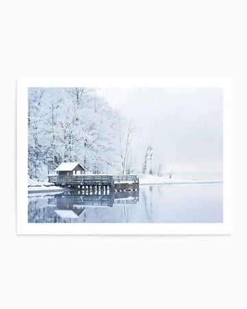Framed photography art print capturing a serene winter landscape with a small wooden boathouse on a pier over a calm lake, printed on fine art paper. The scene is dominated by snow-covered trees lining the far bank, their branches heavily laden with white frost against a soft, overcast sky. The lake's surface reflects the trees and the boathouse, creating a mirror-like effect with subtle ripples. The overall mood is peaceful and tranquil, with cool tones of white, pale blue, and grey.