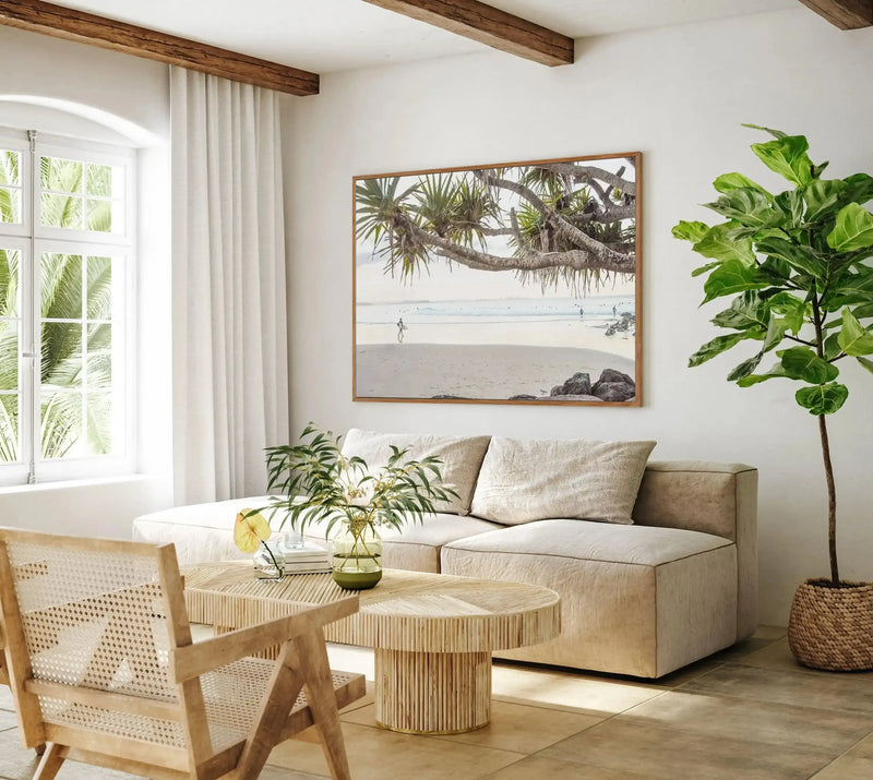 Lifestyle shot of a bright living room with a large framed canvas print of a beach scene displayed above a cream sectional sofa. The print, titled "Last Surf, Snapper Rocks," is a photograph with a natural wood frame and depicts a sandy beach with a person carrying a surfboard walking towards the ocean. The room features a large arched window on the left with views of palm trees, a light wood coffee table with a reeded texture, a woven chair, and a tall fiddle-leaf fig plant in a woven basket on the right.