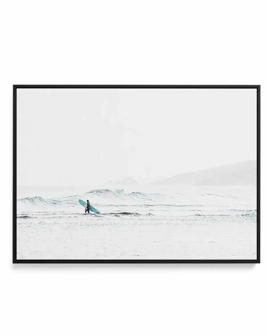 Framed canvas art print featuring a minimalist black and white photograph of a surfer in a teal wetsuit walking into grey, choppy ocean waves on an overcast beach. The composition is horizontally oriented with vast sandy beach in the foreground, rolling waves in the middle ground, and misty mountains visible in the hazy background. The photograph has a cool, muted colour palette with soft, diffused lighting. The canvas is mounted in a black frame with a white border.