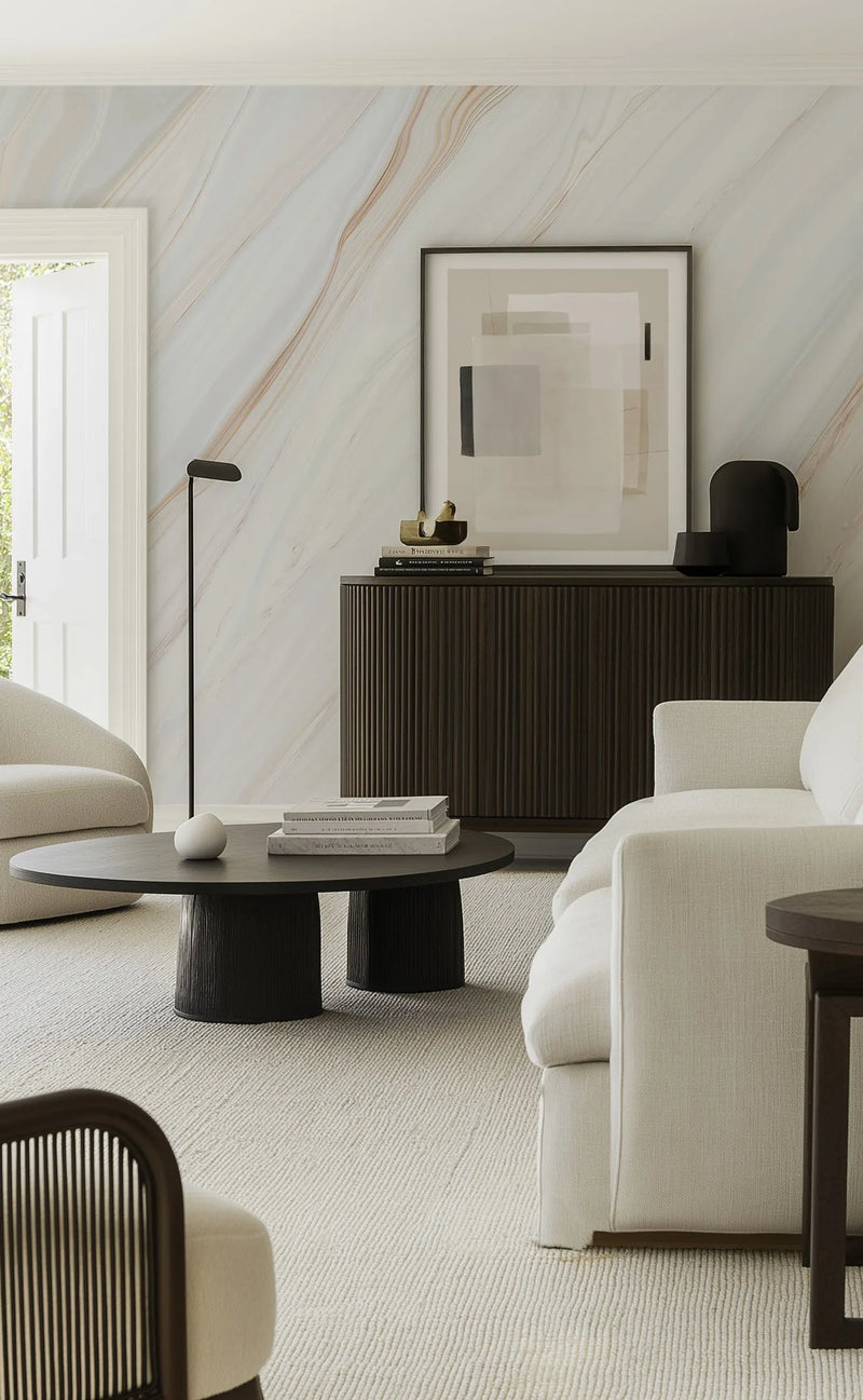 Lifestyle shot of a minimalist living room featuring the Layers Of Earth Wallpaper. The wallpaper features a repeating pattern of soft, flowing marble-like veins in muted shades of cream, pale grey, and warm beige, creating a serene and elegant backdrop. The wallpaper is visible on the wall behind a dark wood fluted console table topped with books and decorative objects, and an abstract framed art print. A cream sofa and a dark wood coffee table are in the foreground.