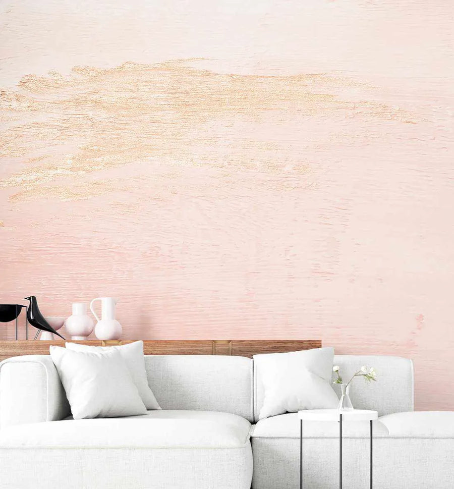 Peel and stick wallpaper featuring a textured, abstract design in soft blush pink with horizontal strokes of metallic gold, displayed on the wall behind a white sectional sofa in a modern living room. The wallpaper has a brushed, painterly quality. On a wooden shelf above the sofa are decorative white vases and a black bird figurine.
