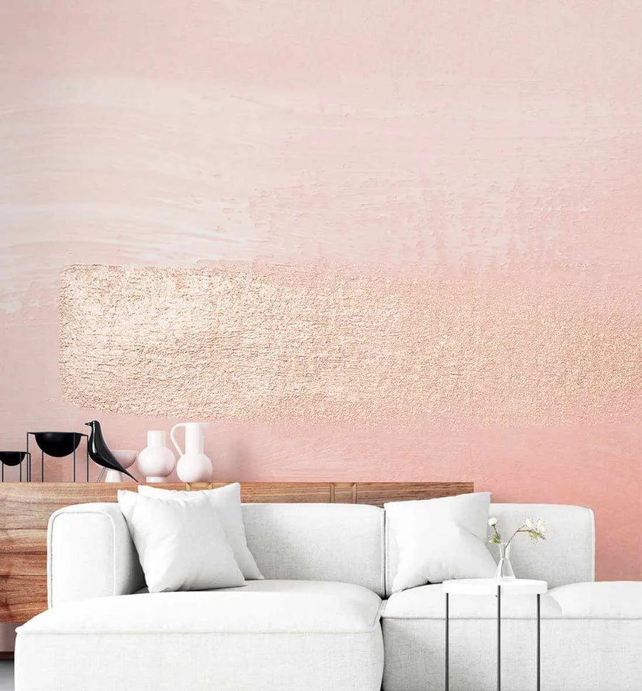 "Peel and stick wallpaper mural featuring an abstract design with horizontal bands of textured blush pink and pale rose, with a central rectangular panel of highly textured, almost granular, light beige. The wallpaper is displayed above a white sectional sofa with white throw pillows and a wooden console table with black and white decorative objects in a minimalist living room."