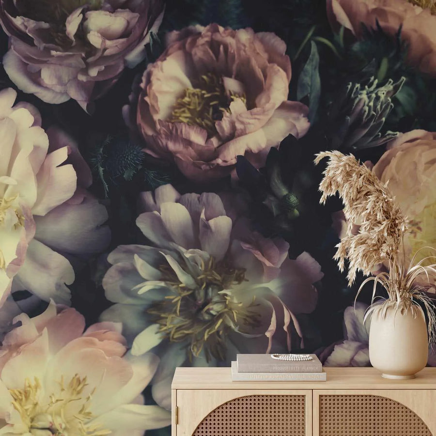 Peel and stick wallpaper featuring a dramatic, dark floral design with large-scale peony blooms in shades of dusty rose, deep plum, and creamy white against a dark, moody background. The wallpaper is displayed on a wall above a light wood console table with woven cane details. On the table, a tall cream vase holds dried pampas grass, and two books are stacked with a beaded bracelet on top.