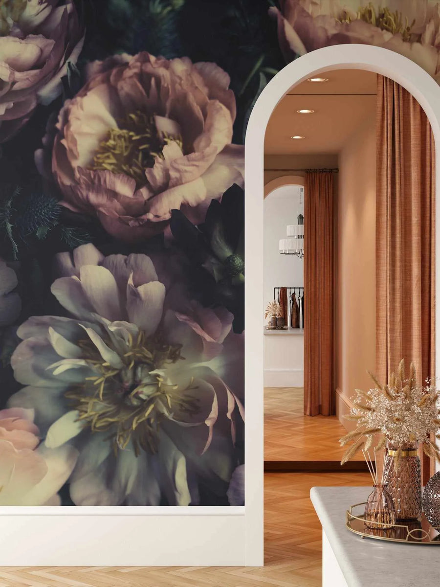 Retail Space:
Le Jardin peel and stick wallpaper mural featuring large-scale, dark and moody peonies in shades of dusty rose, deep plum, and soft cream with visible brushstroke texture. The wallpaper covers a wall in a retail space, with a white arched doorway leading to another room with warm terracotta curtains and a display of dried pampas grass on a white counter.