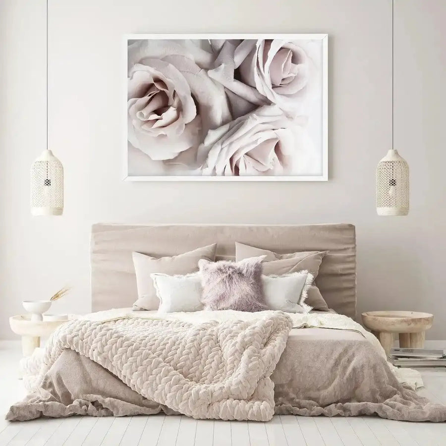 Framed art print displaying a close-up photograph of soft, pale blush and cream-colored roses with layered petals in muted tones. The image has a dreamy, slightly faded quality with subtle shadows emphasizing the delicate texture of the blooms. White frame with white border hung above a beige upholstered bed in a minimalist bedroom setting with neutral bedding and pendant lights on either side.