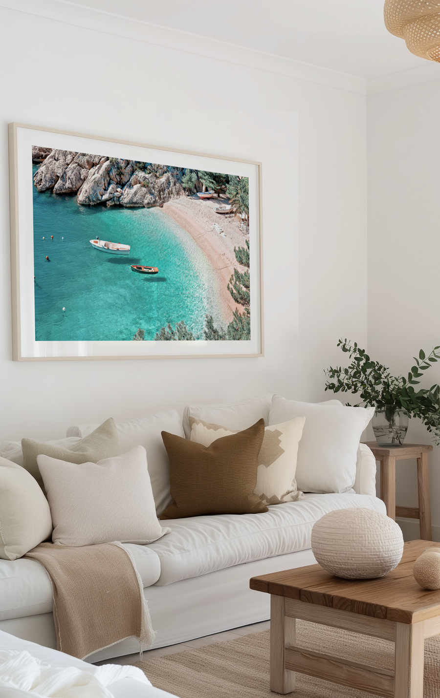 Large framed art print displayed above a white slipcover sofa in a bright, minimalist living room. The print is a vibrant aerial photograph of a secluded beach with turquoise water, a sandy shore, and rocky cliffs, framed in natural wood with a white border. The room features neutral-toned pillows, a beige throw, a light wood coffee table with decorative spheres, and a vase of eucalyptus branches.
