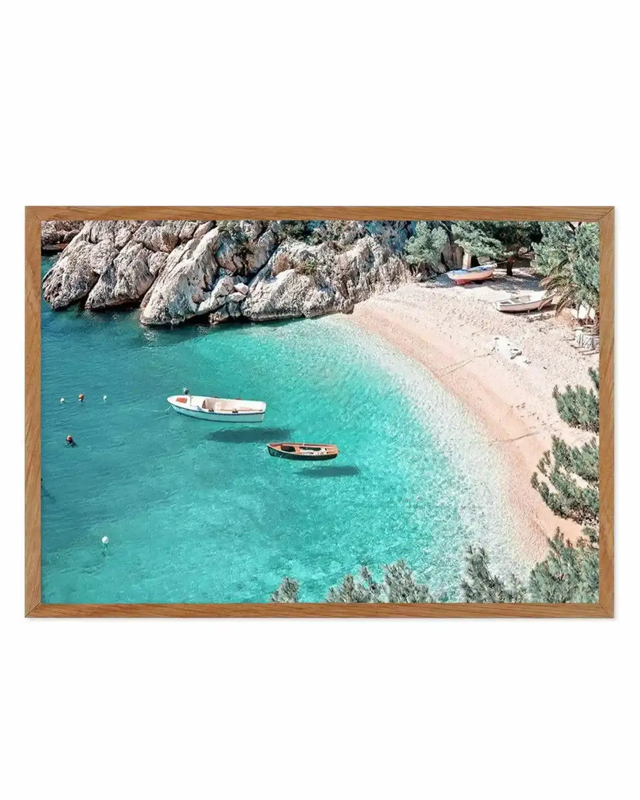 Framed art print depicting an aerial coastal scene with turquoise water, sandy beach, and dramatic light gray rocky cliffs. Two wooden boats float in the shallow water while swimmers enjoy the cove. Pine trees dot the shoreline. The photograph is set in a natural wood frame with white border.