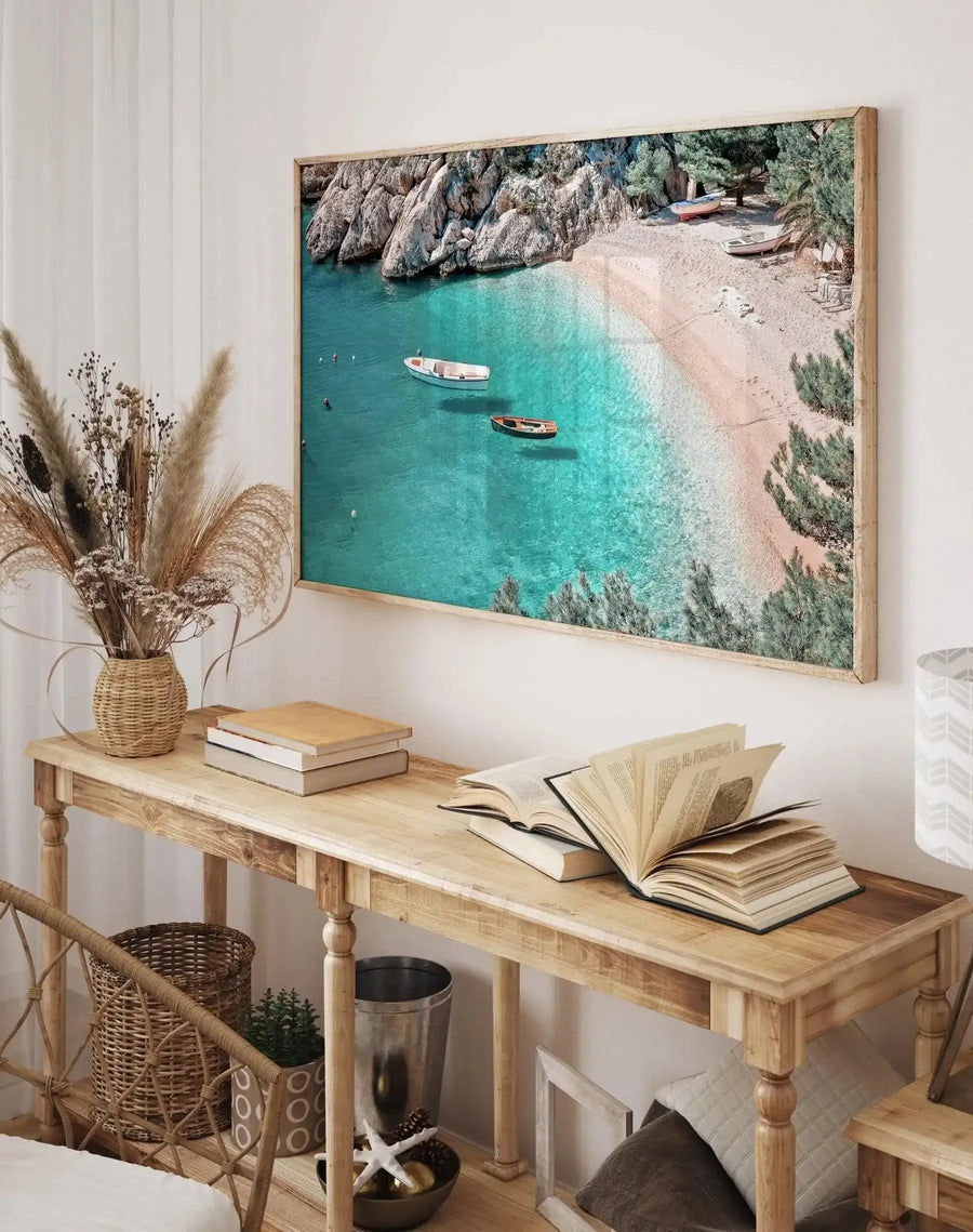 Le Petit Port Art Print displayed on a white wall above a natural wood desk. The print features an aerial photograph of a Mediterranean coastal cove with turquoise water, two small fishing boats, a sandy beach, dramatic white limestone cliffs, and Mediterranean vegetation. The print has a light natural wood frame. Below the artwork, a wooden desk holds open books, a wicker vase with dried pampas grass, stacked books, and decorative objects including a starfish and small bowl.