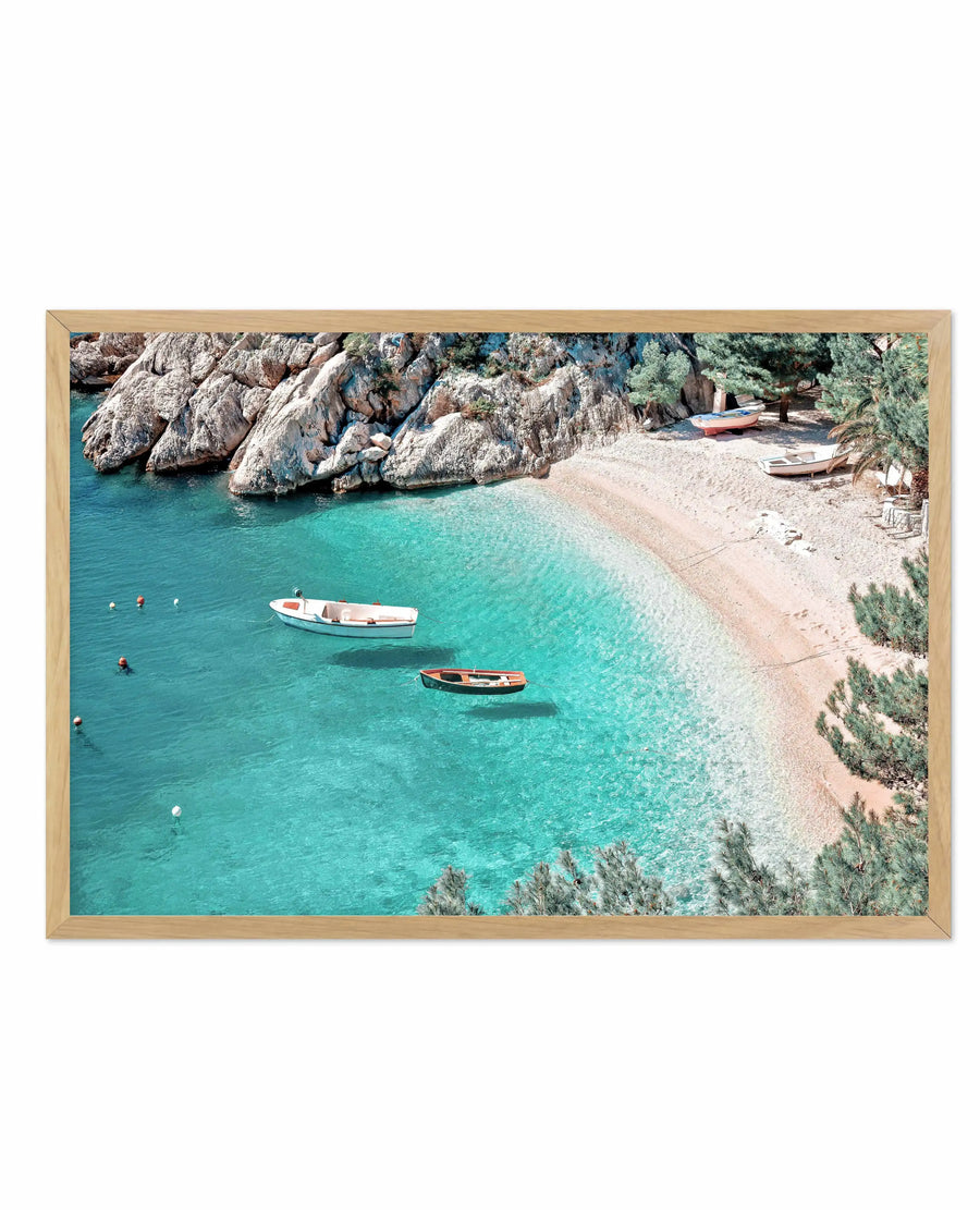 Framed art print depicting an aerial view of a Mediterranean coastal cove with crystal-clear turquoise water, two fishing boats anchored near a sandy beach, rocky limestone cliffs covered in green vegetation, swimmers in the water, and additional boats beached on shore. Photograph with natural colors in warm sand tones, cool aqua blue water, and grey-white rock formations. Natural wood frame with horizontal orientation.