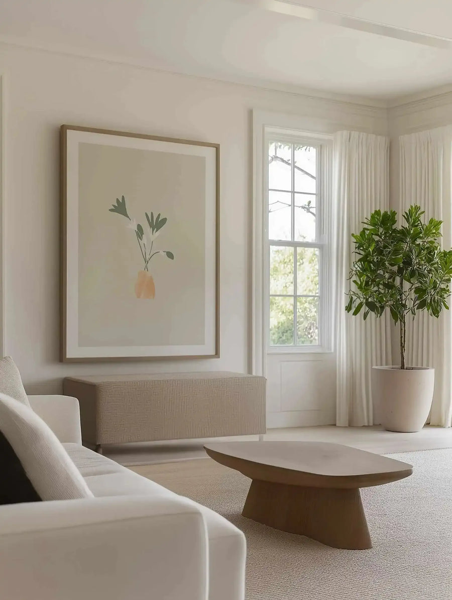 Lifestyle shot of a minimalist living room featuring a framed art print above a textured beige console table. The print, "Le Posy III," depicts a minimalist illustration of three stylized flowers with dark green leaves and white petals in a small, abstract orange vase, set against a pale cream background, with a natural wood frame and white border. A white sofa is partially visible in the foreground, and a large potted plant stands next to a window with white curtains.