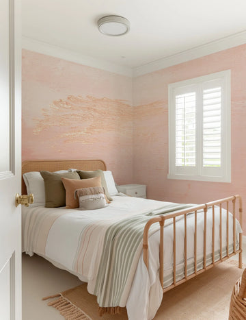 "Peel and stick wallpaper featuring an abstract, textured design in soft blush pink with horizontal strokes of metallic gold, creating a subtle, organic pattern on the walls of a cozy bedroom. The wallpaper is visible behind a rattan headboard and bed dressed in white and striped bedding, with a small white nightstand and a window with white plantation shutters."