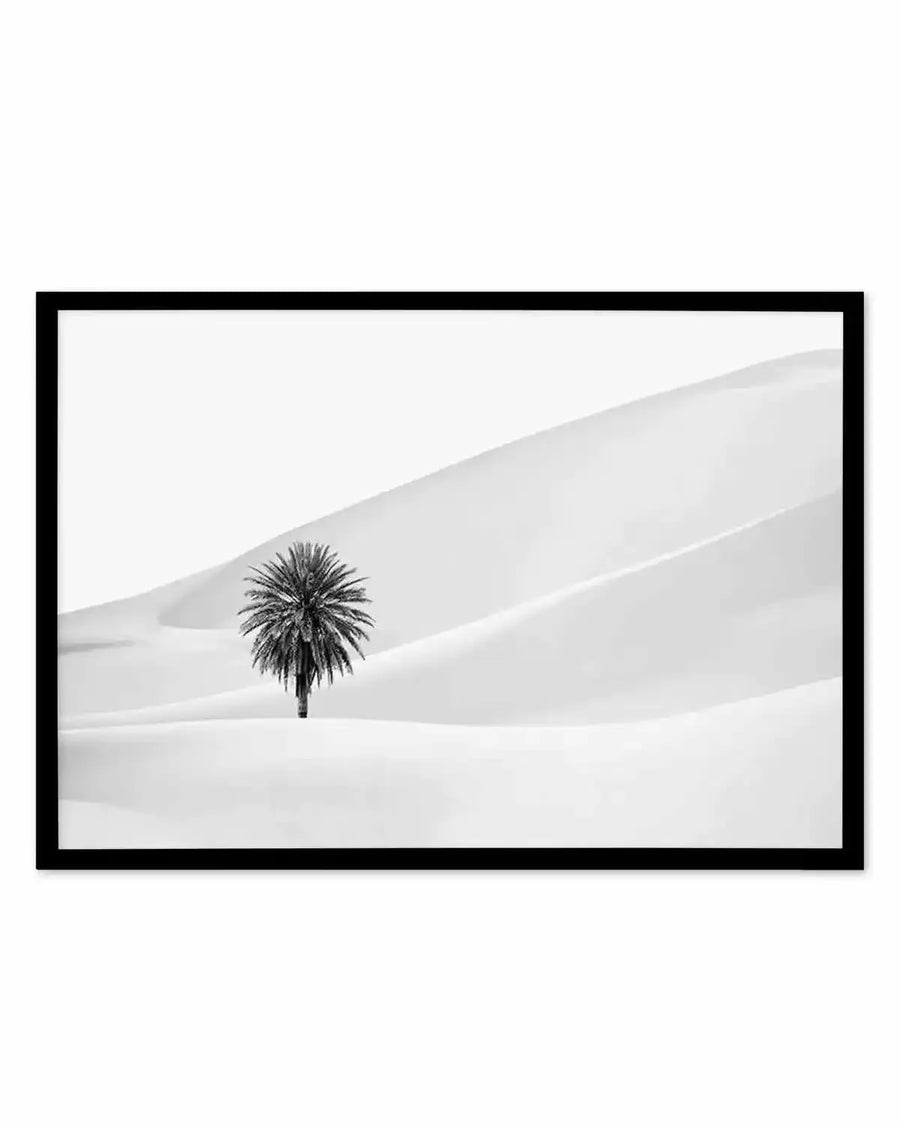 Framed black and white photograph of a solitary palm tree in a vast desert landscape, with a black frame and white border. The composition features a single palm tree with dark, dense fronds and a slender trunk, standing in the middle of gently undulating sand dunes. The sky is a bright, stark white, creating a high-contrast, minimalist scene.