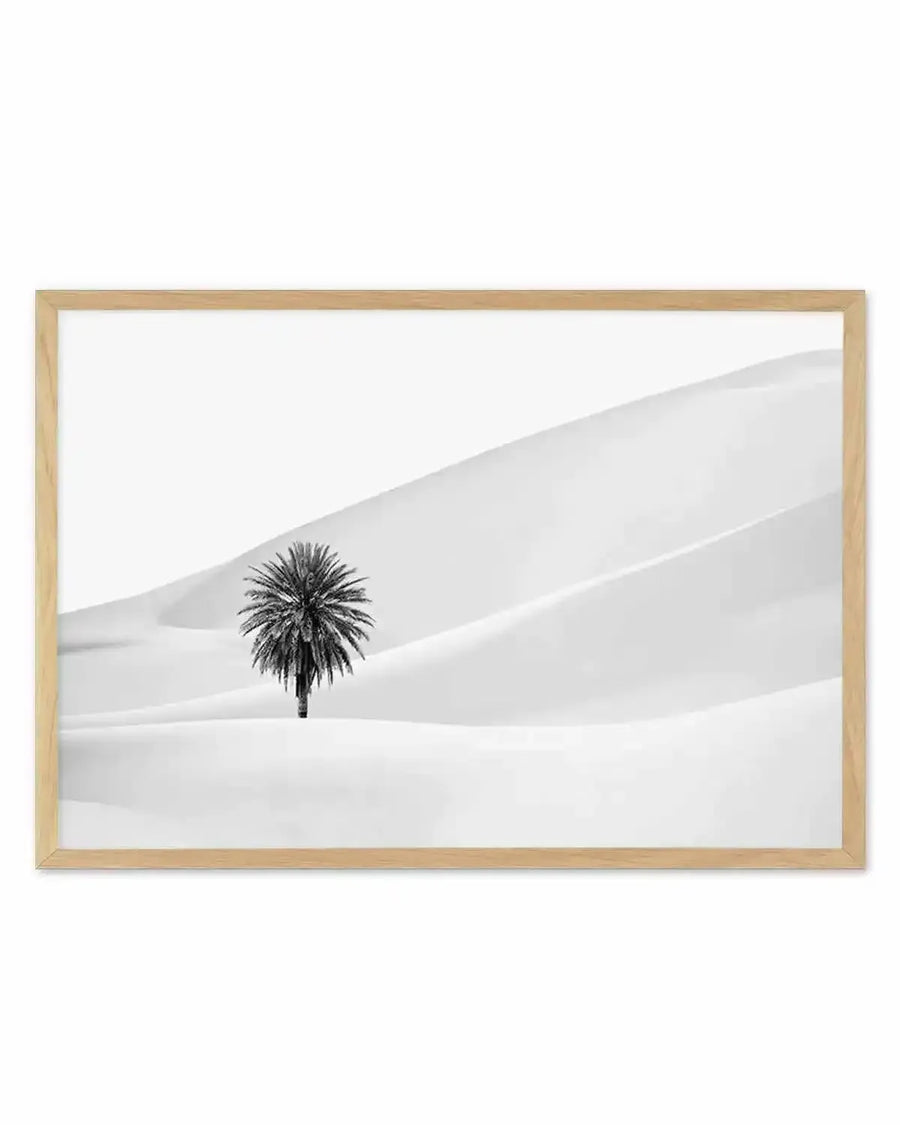 Framed black and white photograph of a lone palm tree in a vast desert landscape with natural wood frame and white border. The composition features a single palm tree with dark foliage and trunk standing in the middle of undulating white sand dunes under a bright white sky.