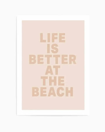 Art print featuring the phrase "LIFE IS BETTER AT THE BEACH" in bold, sans-serif, light beige capital letters on a soft blush pink background, displayed with a white border.