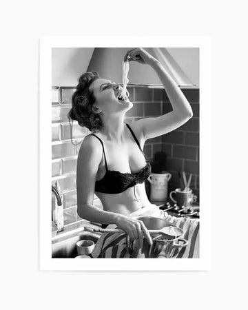 Framed black and white photograph of a woman in a kitchen, sitting on a counter next to a sink, wearing a black bra, and playfully eating a long strand of spaghetti with her mouth wide open. Her hair is styled in retro curls, and she looks up at the pasta with an expression of joy. The background features subway tiles and kitchen appliances, with a pot and striped towel visible in the foreground.