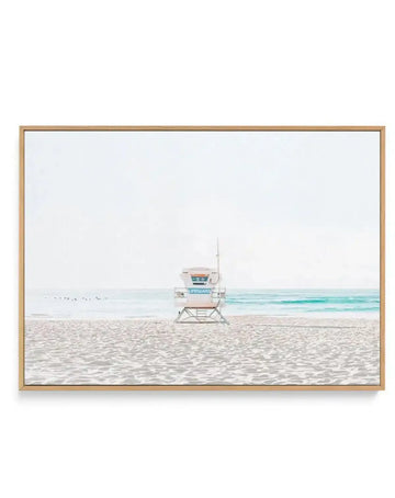 Framed canvas art print in natural wood frame featuring a photograph of a white and turquoise lifeguard tower standing alone on sandy beach with calm turquoise ocean and overcast sky in background. The image has a soft, minimalist aesthetic with neutral sand tones, pale sky, and clear water. The canvas is mounted in a light wood frame with white border.