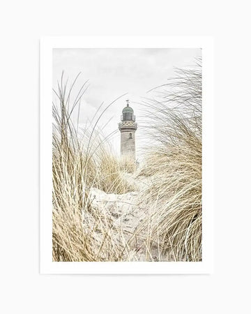 Framed black and white photograph of a coastal scene with a lighthouse and sand dunes, with a white border. The composition features a tall, slender lighthouse with a dark dome, partially obscured by tall, dry grass and sand dunes in the foreground. The sky is overcast and bright.