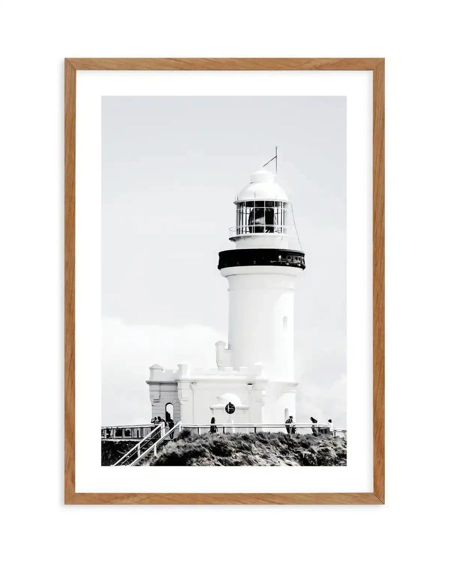 Framed black and white photograph of a lighthouse on a hill with a natural wood frame and white border. The composition features a tall, white lighthouse with a black base and a glass-enclosed lamp room, set against a bright, overcast sky. Below the lighthouse, a white building with arched doorways and a railing is visible, with people standing on a grassy, textured hill.