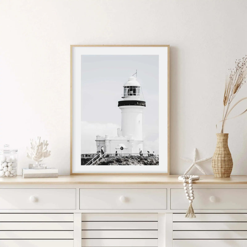 Lifestyle shot of a framed black and white photograph of a lighthouse displayed on a white console table in a bright room. The print features the Byron Bay lighthouse in a monochrome palette with a natural wood frame and white border. The console table is adorned with coastal-themed decor including a glass jar filled with white pebbles, a white coral sculpture, a white starfish, a woven vase with dried wheat, and a string of white wooden beads.