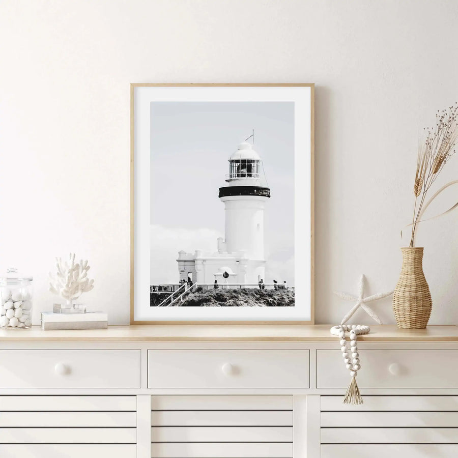Lifestyle shot of a framed black and white photograph of a lighthouse displayed on a white console table in a bright room. The print features the Byron Bay lighthouse in a monochrome palette with a natural wood frame and white border. The console table is adorned with coastal-themed decor including a glass jar filled with white pebbles, a white coral sculpture, a white starfish, a woven vase with dried wheat, and a string of white wooden beads.