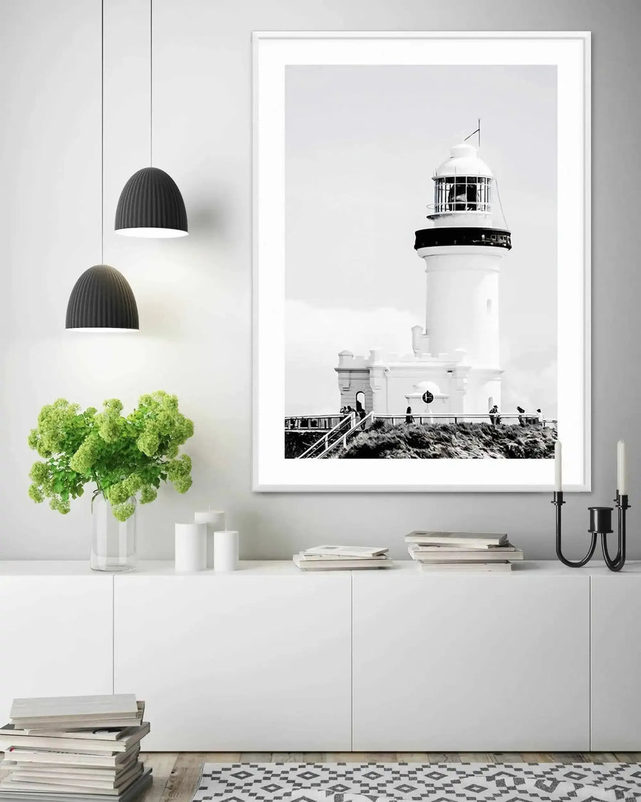 Large framed black and white photograph of a lighthouse displayed above a white console table in a minimalist living room. The print features the white lighthouse of Byron Bay on a cliff overlooking the ocean with people walking on a path below. The room includes two black pendant lights, a vase of green hydrangeas, white candles, stacks of books, and a black and white patterned rug.