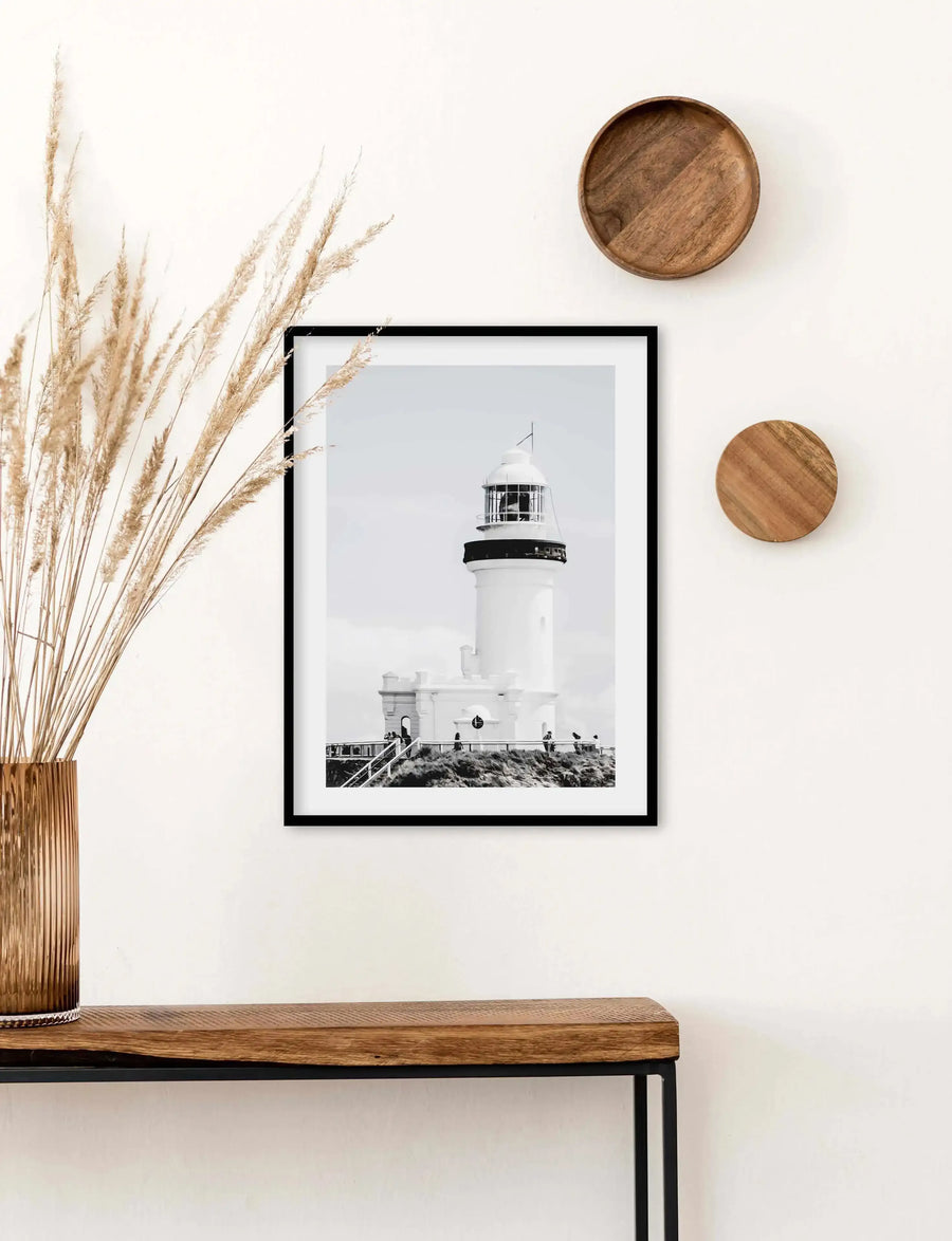 Lifestyle shot of a framed black and white photograph of a lighthouse displayed above a wooden console table in a minimalist room. The print features a white lighthouse with a black top against a bright white sky, with a low stone wall and grassy hill in the foreground. The print is in a black frame with a white border. The room includes a tall copper vase with dried pampas grass, and two round wooden decorative pieces on the wall.