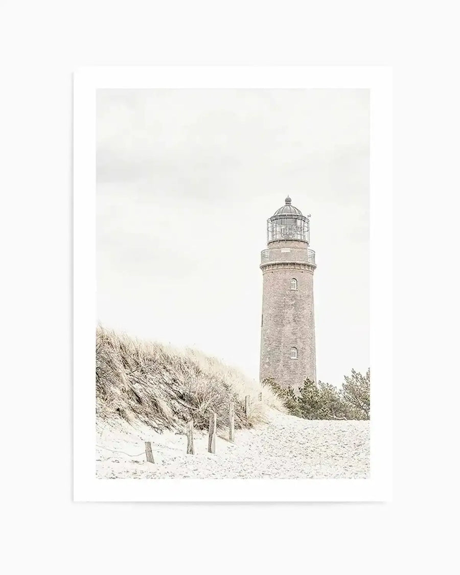 Framed black and white photograph of a coastal beach scene with a tall lighthouse and sand dunes, with natural wood frame and white border. The composition features a brick lighthouse on the right, with sandy dunes and dry grass on the left, under an overcast sky.