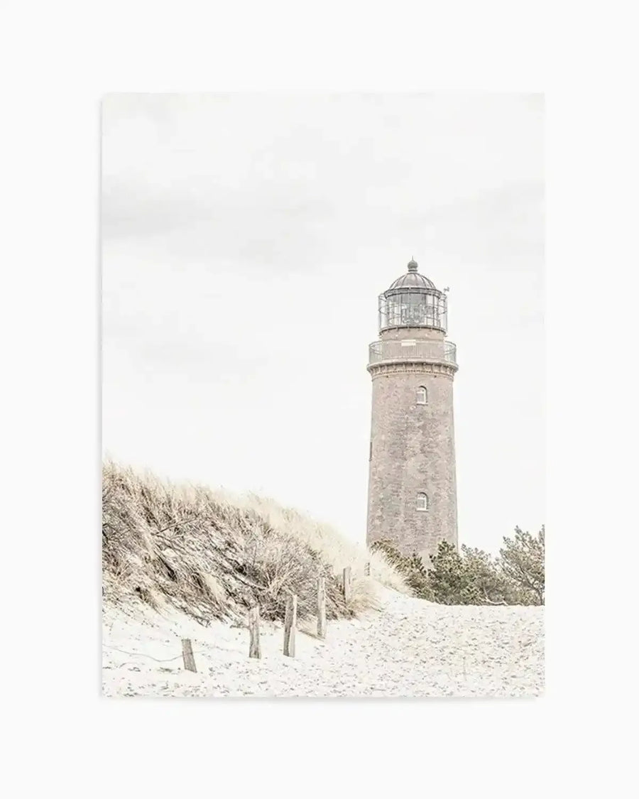 Framed black and white photograph of a coastal scene with a lighthouse and sand dunes, with natural wood frame and white border. The composition features a tall, slender lighthouse on the right, with sandy dunes and sparse, dry grasses on the left, under an overcast sky.