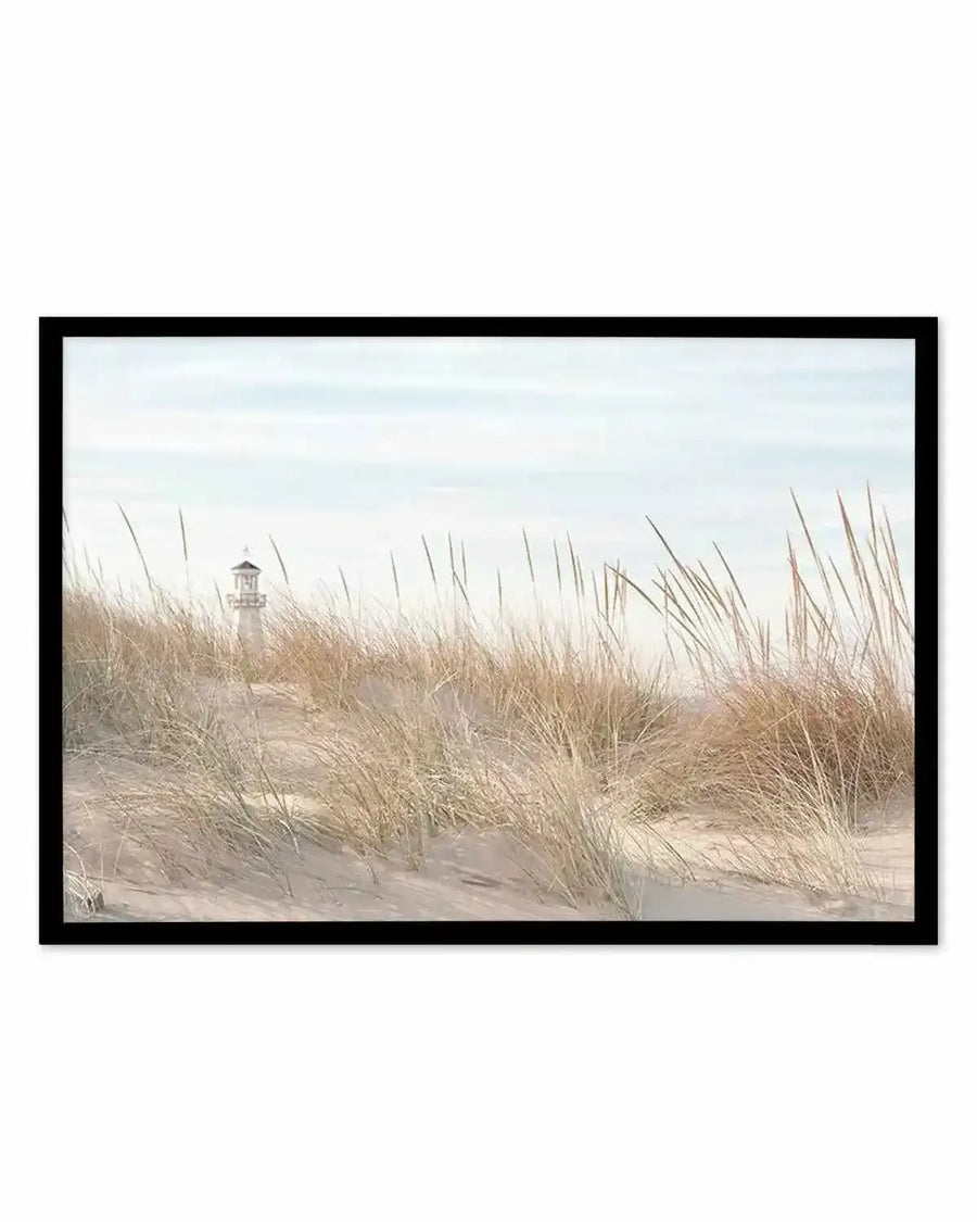 Framed photograph of a coastal beach scene with a black frame and white border. The composition features a distant white lighthouse peeking over sandy dunes covered in dry, light brown and golden grasses under a pale blue, slightly overcast sky.