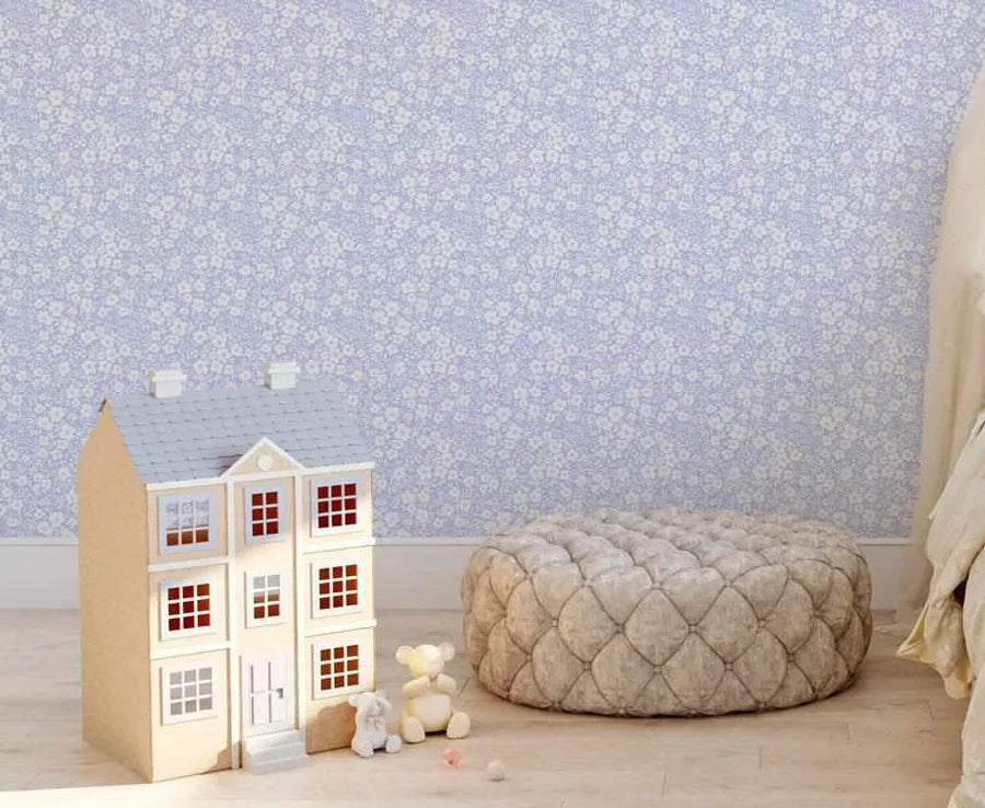 Lifestyle shot of a child's room featuring Lilac Fleur wallpaper. The wallpaper has a repeating pattern of small, delicate white flowers with subtle green leaves on a soft lilac background, creating a gentle, floral design. A light brown wooden dollhouse with a grey roof and white window frames sits on a light wooden floor to the left, with two small teddy bears and pink beads nearby. A large, round, tufted beige ottoman is on the floor to the right, next to a cream-colored bed with a duvet.