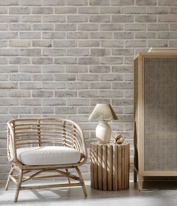 "Lime Washed Brick Wallpaper applied to a wall in a bright, minimalist living room featuring a rattan armchair with a white cushion, a wooden side table with a lamp, and a wooden cabinet. The wallpaper has a repeating pattern of white and light grey bricks with visible texture and subtle variations in tone, creating a rustic yet clean aesthetic."