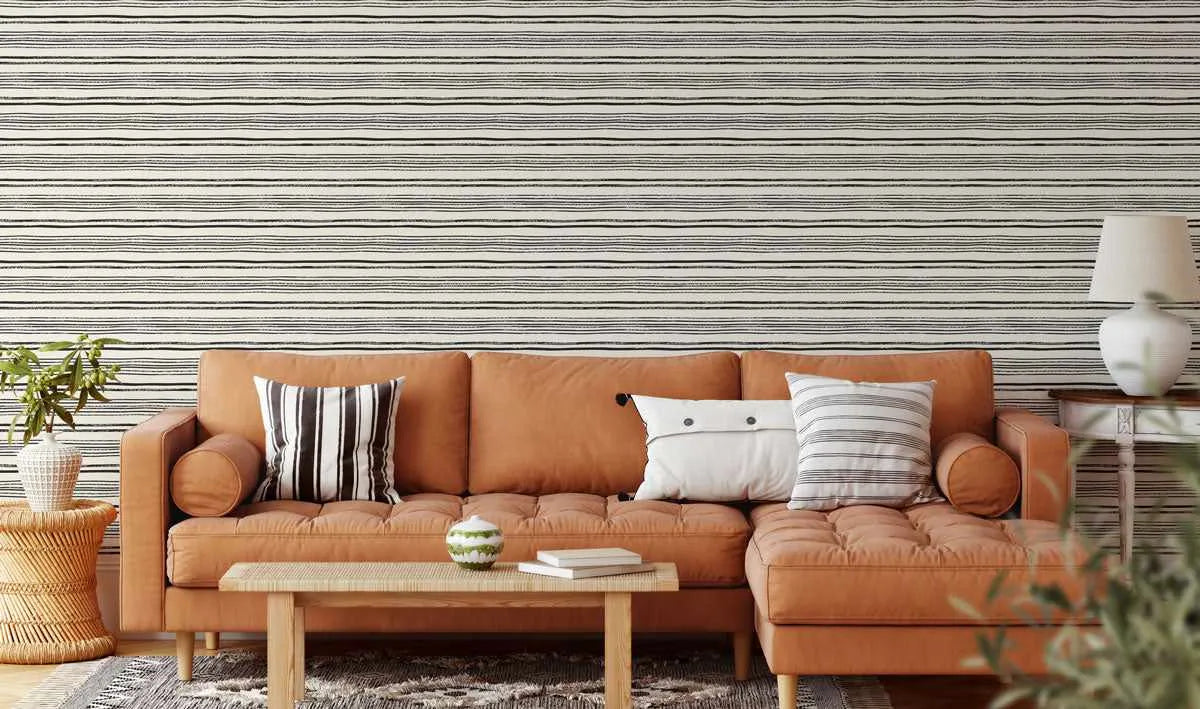 Lifestyle shot of a living room featuring a wall covered in wallpaper with a repeating pattern of thin, hand-drawn horizontal black stripes on a cream background. A rust-orange sectional sofa with tufted cushions is placed against the wall, adorned with various throw pillows including a black and white striped one and a white one with button details. A light wood coffee table sits in front of the sofa, and a white table lamp on a small side table is visible on the right.