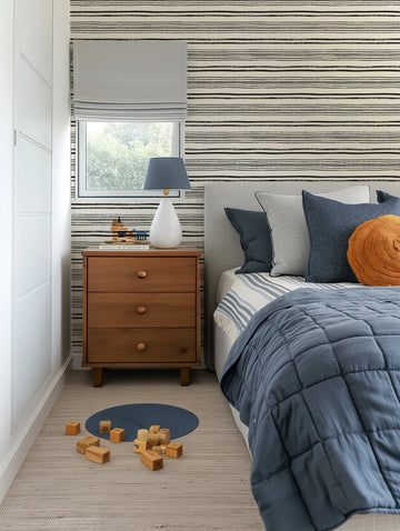 Lifestyle shot of a child's bedroom featuring a wallpaper with a repeating pattern of hand-drawn horizontal stripes in black on a cream background. The stripes vary in thickness and texture, some appearing solid and others with a distressed, brushstroke quality. The wallpaper covers the wall behind a bed with a light grey upholstered headboard and a wooden nightstand. The room includes a window with a light grey roman blind, a blue quilted duvet, and wooden toys on a light-coloured rug.