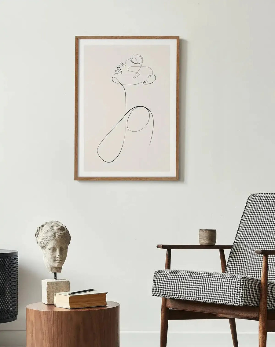 Large framed art print displayed on a white wall above a wooden side table in a minimalist living room. The print features a single continuous line drawing of a woman's face and upper body, looking upwards, in black on a cream background, with a natural wood frame and white border. The room includes a stone bust, a book, and a black and white houndstooth armchair.
