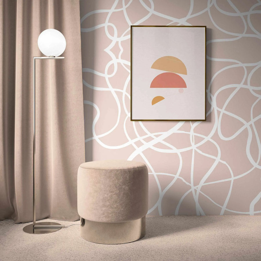 Lifestyle shot of a room featuring peel and stick wallpaper with a repeating pattern of abstract, free-flowing white lines on a nude pink background, creating a dynamic, intertwined design. The wallpaper covers the wall behind a tall, minimalist floor lamp with a spherical white shade and a round, plush nude pink ottoman with a metallic base. The room has a light beige carpet and a floor-length nude pink curtain on the left.