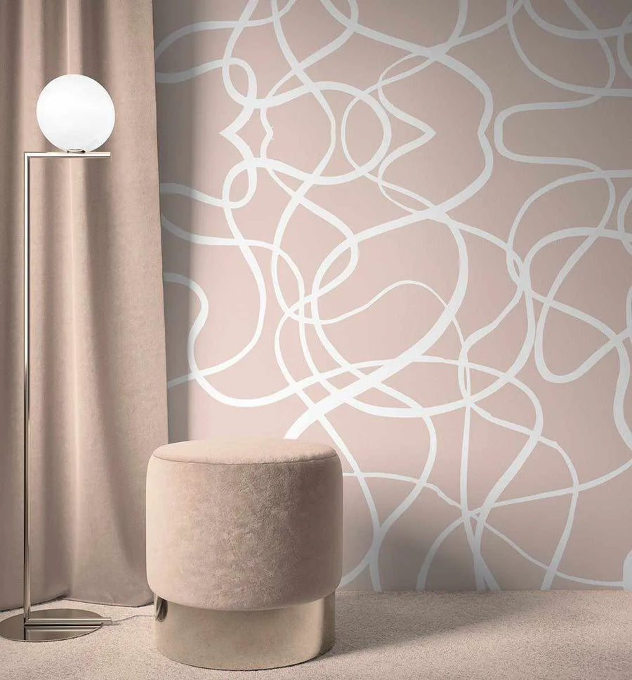 Peel and stick wallpaper featuring a repeating pattern of abstract, free-flowing white lines on a nude pink background, displayed on a wall in a minimalist living room. The room includes a tall silver floor lamp with a spherical white shade, a plush nude pink velvet stool with a metallic base, and a matching nude pink curtain.