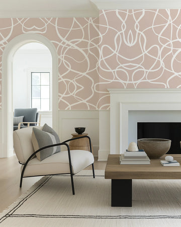 Lifestyle shot of a living room featuring a peel and stick wallpaper with a repeating pattern of abstract, free-flowing white lines on a nude pink background. The wallpaper is applied to the walls of a living room with white wainscoting and crown molding, an arched doorway leading to another room, a white fireplace, a cream armchair with black metal arms, a wooden coffee table, and a light-colored rug with black stripes.