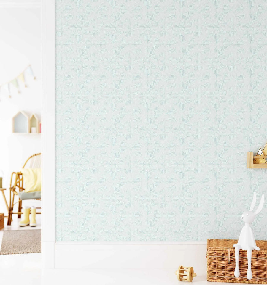 Lifestyle shot of a child's room featuring a wall covered in peel and stick wallpaper with a repeating pattern of concentric arches in a soft aqua blue on a white background. The pattern creates a subtle scalloped or fan-like motif. The room includes a wicker chair with a yellow cushion, a small rug, and a white bunny figurine sitting on a woven basket.