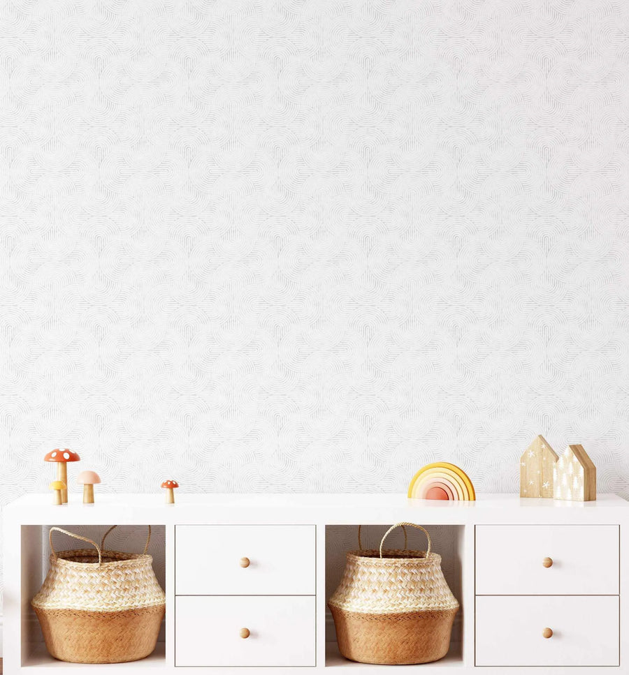 Lifestyle shot of a children's room featuring Little Arches in Grey wallpaper. The wallpaper has a repeating pattern of interlocking semi-circles in a light grey on a white background, creating a subtle, textured geometric design. The wallpaper is visible behind a white storage unit with open shelves and drawers, displaying woven baskets and wooden toys including mushrooms, a rainbow, and houses.