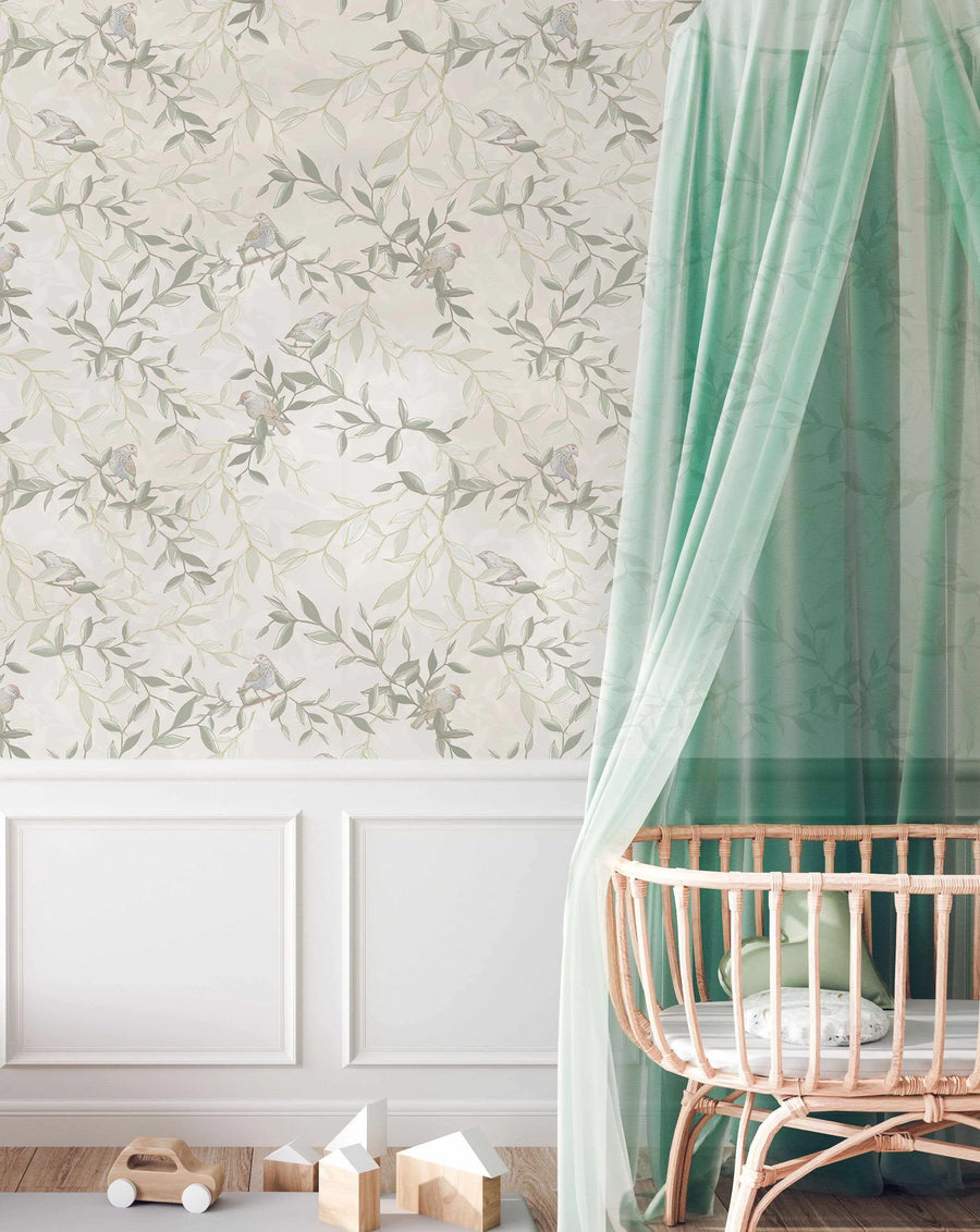 Nursery:
"Peel and stick wallpaper featuring a repeating pattern of hand-painted birds perched on leafy branches in muted sage green and light olive on a soft cream background, with a loose, organic watercolour quality. The wallpaper is displayed in a nursery above white wainscoting, with a light rattan crib and sheer mint green canopy on the right, and wooden toy blocks and a wooden toy car on the floor in the foreground."