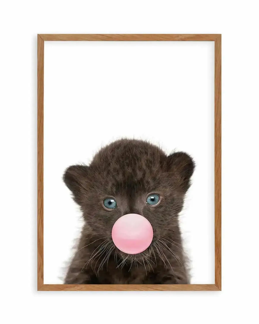 Art print in natural wood frame featuring a young black leopard cub with striking blue-green eyes blowing a large bubble gum bubble in soft pink. The cub's dark fur is rendered in detailed, photorealistic style against a clean white background. The composition is centered and vertical, with the cub's face filling most of the frame. White border surrounds the artwork within the light wood frame.