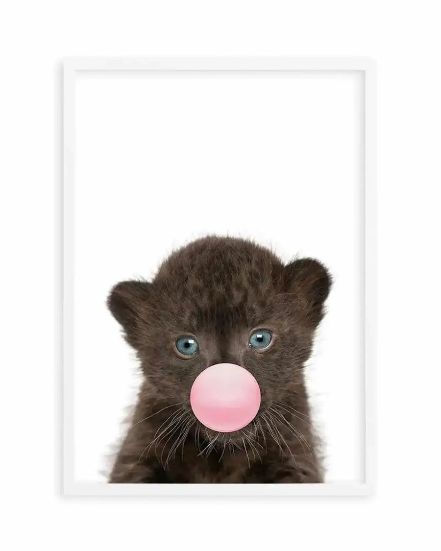 A black leopard cub with striking blue-green eyes gazes directly forward against a white background, blowing a large glossy bubble gum bubble in soft pink. The photograph-style art print is framed in white with a white border, capturing fine details of the cub's dark fur texture and delicate whiskers. The composition centers the young animal's face in a portrait style, creating a playful and whimsical mood through the contrast of the wild animal with the childlike act of bubble-blowing.