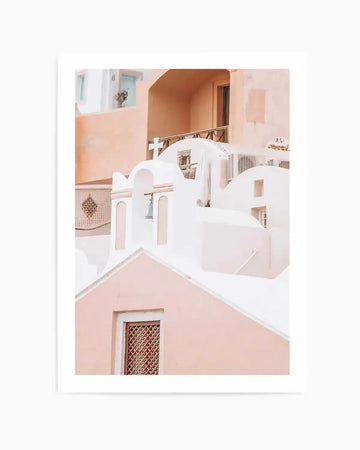 Santorini Art Print framed in white with white border, featuring a photograph of Mediterranean architecture with whitewashed buildings, curved archways, and rounded domed structures. Soft peachy-pink and terracotta tones accent the white buildings. A wooden railing with metal details visible in upper portion. Decorative lattice work and small windows characteristic of Greek island design. Warm, minimalist aesthetic with gentle color palette of cream, dusty rose, and warm tan tones.