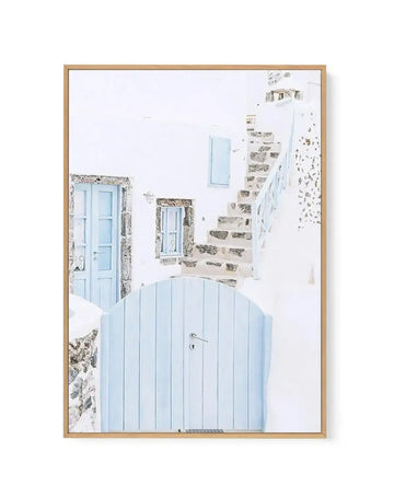 Framed canvas art print featuring a photograph of a white-washed Mediterranean alleyway with light blue wooden gate in foreground, weathered stone walls, blue door and window shutters, and stone staircase receding into background. Photograph has soft, bright, minimalist aesthetic with predominantly white and pale blue tones. Natural wood frame with white border surrounding the image.