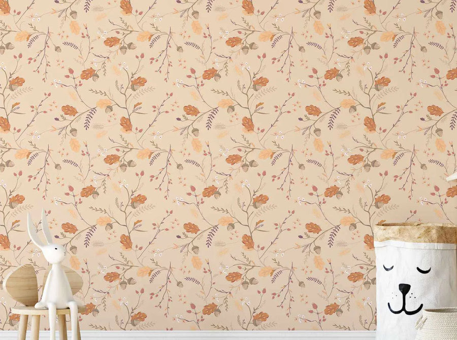 Lifestyle shot featuring Little Boho Garden Wallpaper in a child's room. The wallpaper has a repeating pattern of delicate branches with autumn leaves in shades of burnt orange, golden yellow, and deep red, interspersed with small white berries and acorns, all on a soft beige background. A white rabbit figurine sits on a light wood stool in the foreground, and a white fabric storage basket with a smiling bear face is visible on the right.