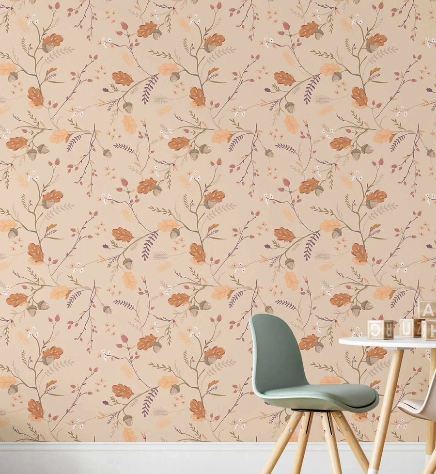Peel and stick wallpaper featuring a repeating pattern of hand-painted autumn foliage on a light beige background. The design includes oak leaves in shades of rust and burnt orange, acorns with light brown caps, delicate white berries, and slender branches with small purple and golden-yellow leaves. The botanical elements have a soft, illustrative quality. In the foreground, a light green chair with wooden legs is pulled up to a small white table with wooden alphabet blocks.