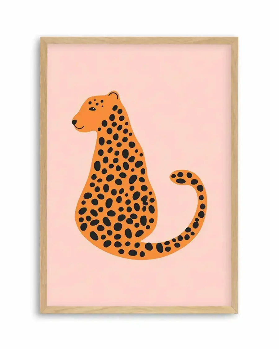 Illustrated art print featuring a stylized cheetah in a seated position, facing left with a gentle smile, against a soft pastel pink background. The cheetah is depicted in a warm orange hue with numerous irregular black spots covering its body and tail. The print is displayed in a natural oak frame with a white border.