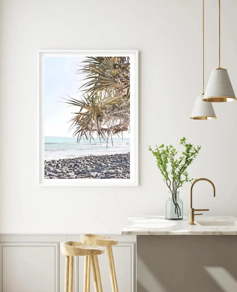 Lifestyle shot of a modern kitchen with a framed art print displayed on a white wall. The print features a vibrant beach scene with a pandanus tree in the foreground, its spiky leaves and exposed roots hanging over a pebbled beach. The ocean in shades of turquoise and blue stretches to the horizon under a clear sky. The room includes two light wood bar stools, a white marble countertop with a gold faucet, a glass vase with green foliage, and two concrete and gold pendant lights.