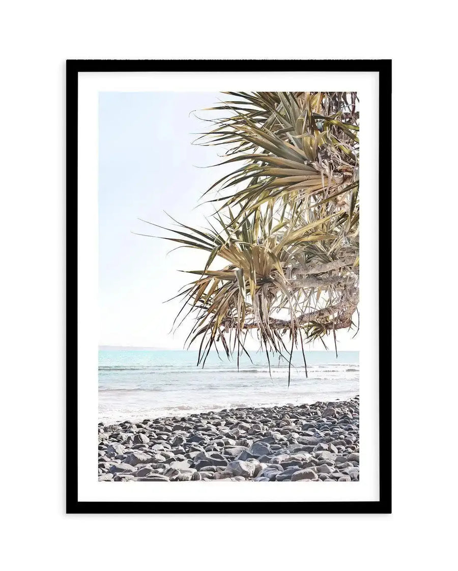 Framed photography art print of a coastal scene with a black frame and white border. The image captures the lower branches of a pandanus tree with long, spiky green and yellow leaves hanging down, obscuring the upper part of the sky. Below the tree, a calm ocean with gentle waves meets a rocky beach composed of smooth, grey stones. The sky is a clear, pale blue.